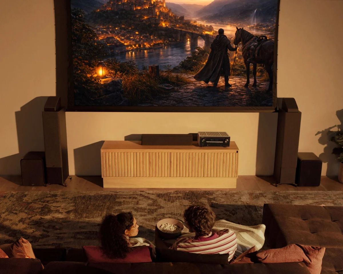 Zwei Personen auf dem Sofa vor der Leinwand, darunter Lautsprecher und Verstärker