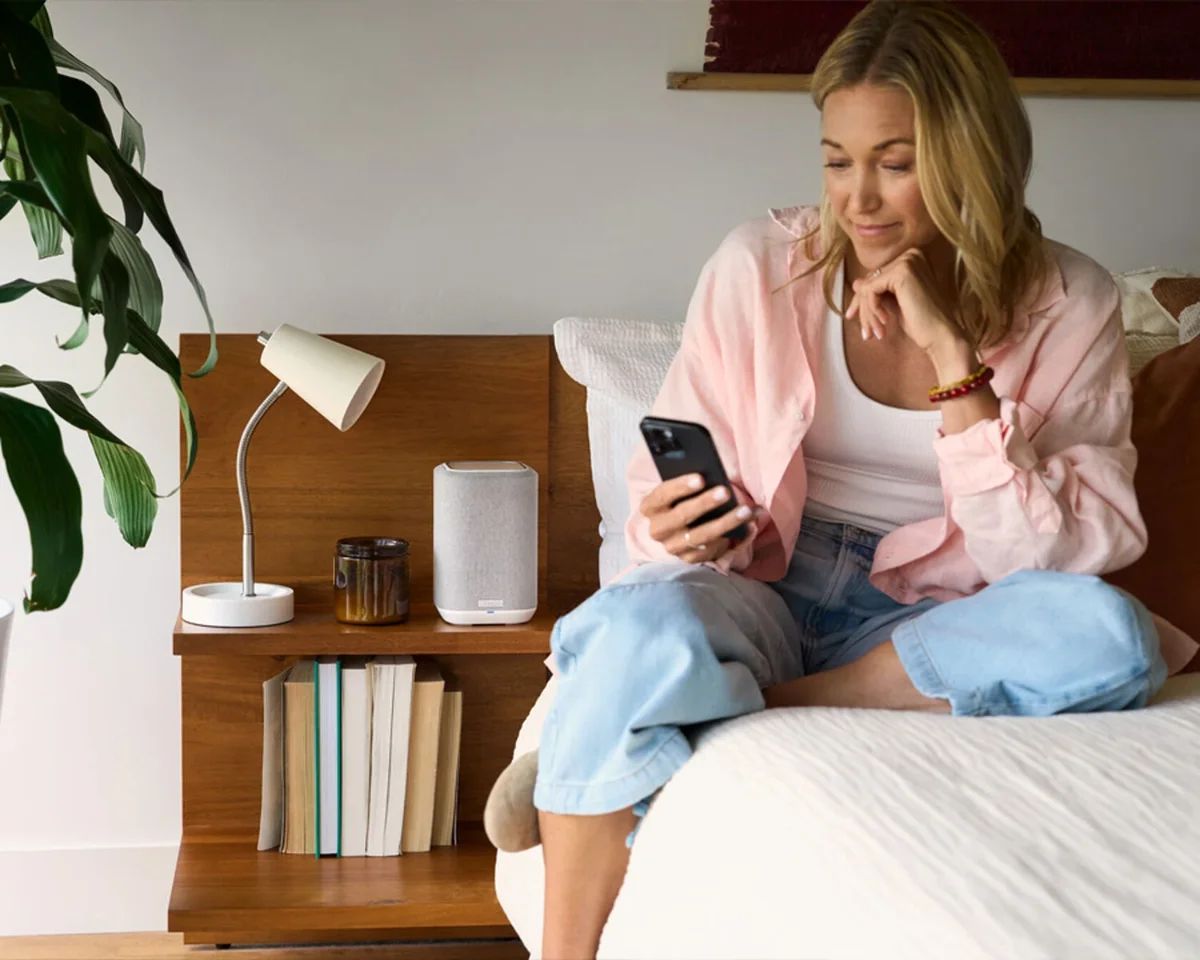 Frau sitzt mit Smartphone auf dem Bett, auf dem Nachttisch der Lautsprecher in Weiß