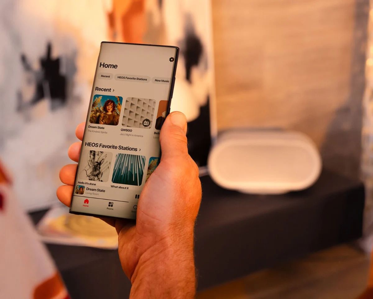 Smartphone mit der HEOS App, dahinter der Lautsprecher in Hellgrau auf einem Sideboard